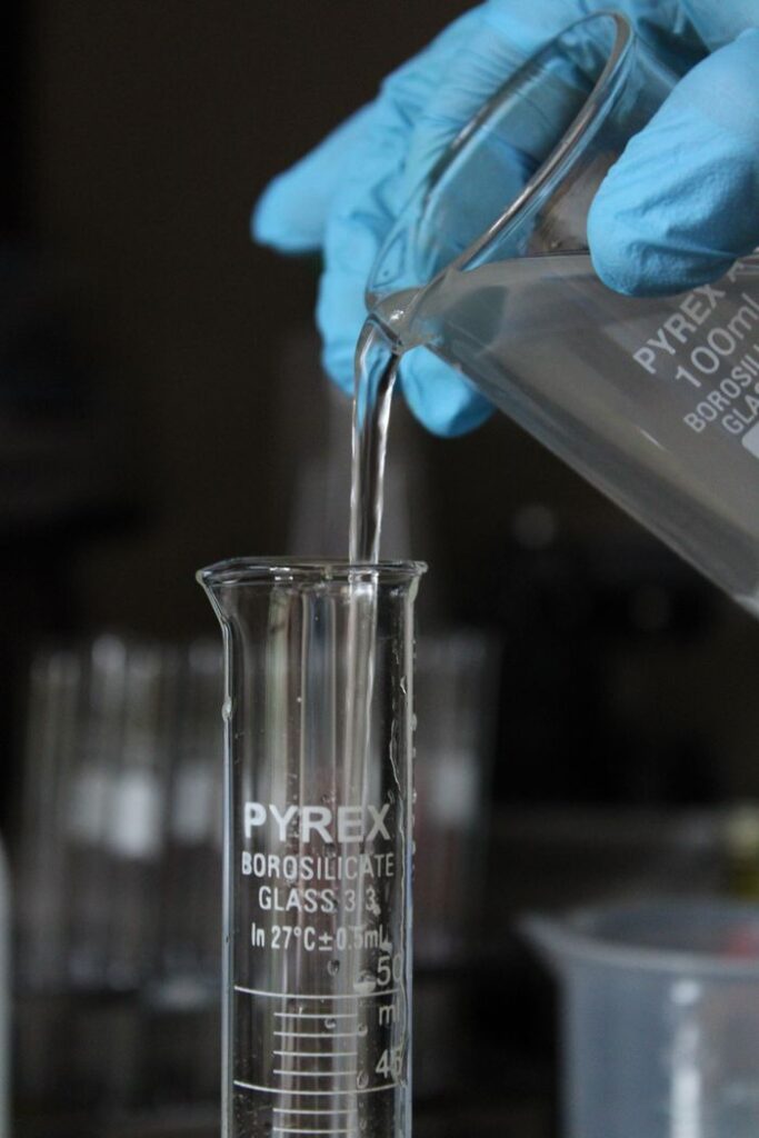 Home Scientist wearing gloves pours liquid into a measuring beaker in a laboratory setting.